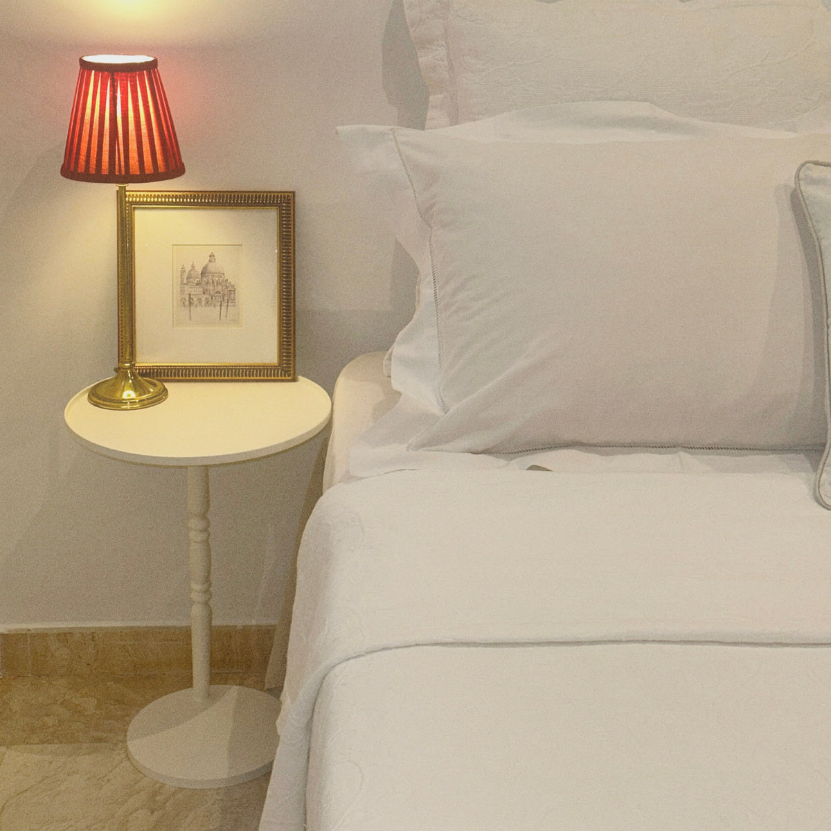 Mesa modelo Gorga con una lámpara y una decoración encima junto a una cama con almohadas